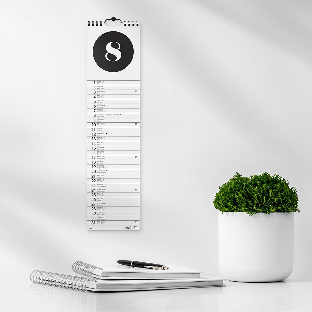 Väggkalender Burde 1768 Simple 2026 med datum och månadsschema, svart och vitt, på vägg med skrivblock och penna framför