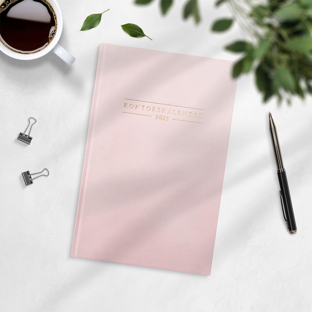 Grön kontorskalender för 2026 med rosa omslag och guldtext, omgiven av penna, klämmor, kopp kaffe och gröna växter