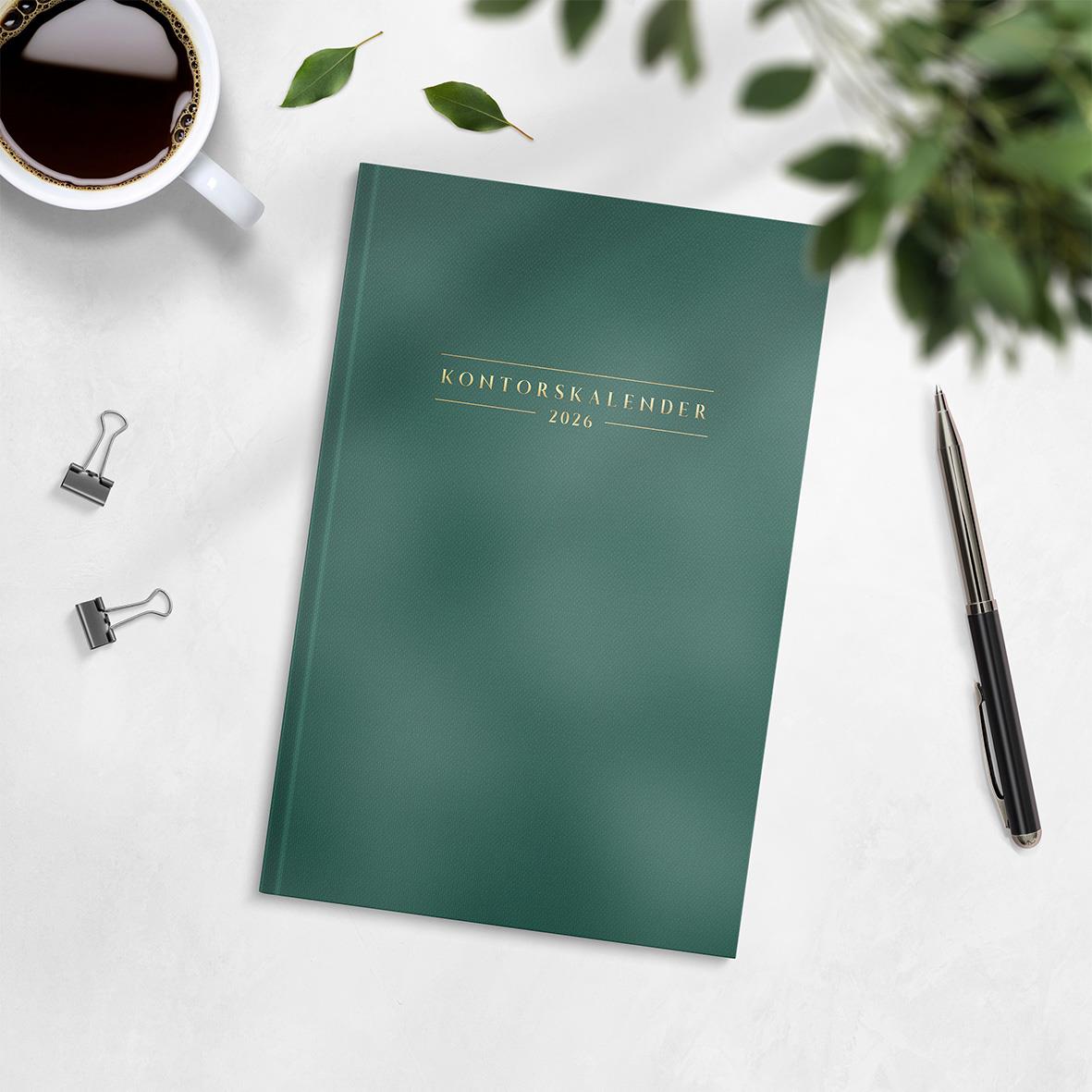 Grön kontorskalender för 2026 med titeln Kontorskalender 2026 på omslaget, omgiven av penna, klämmor, kaffekopp och gröna växter