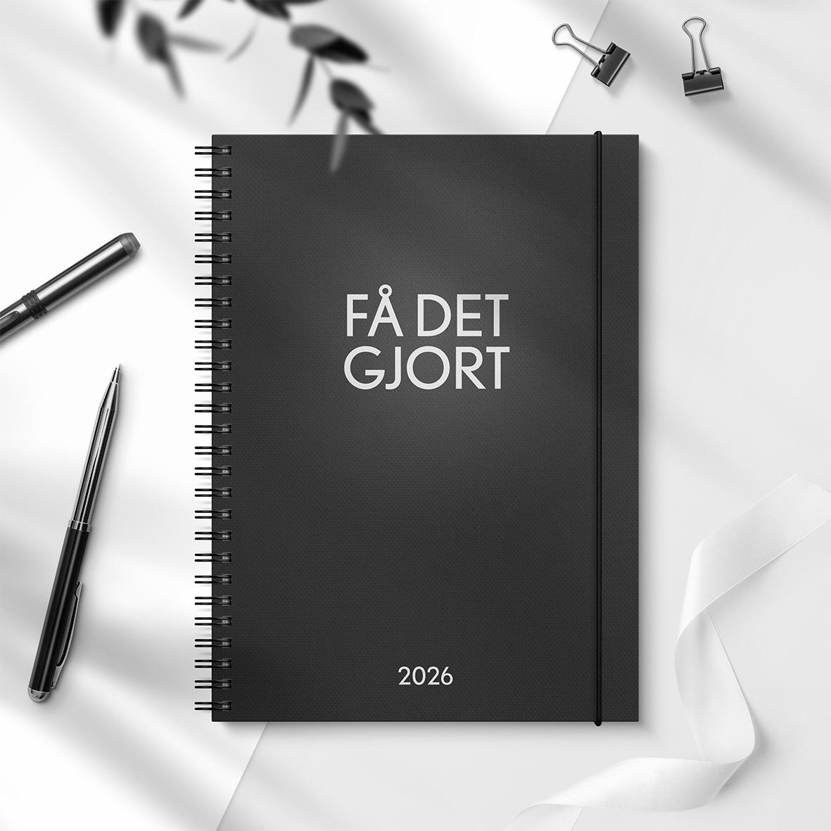 Svart almanacka 2026 med text FÅ DET GJORT på framsidan, omgiven av pennor och kontorsmaterial