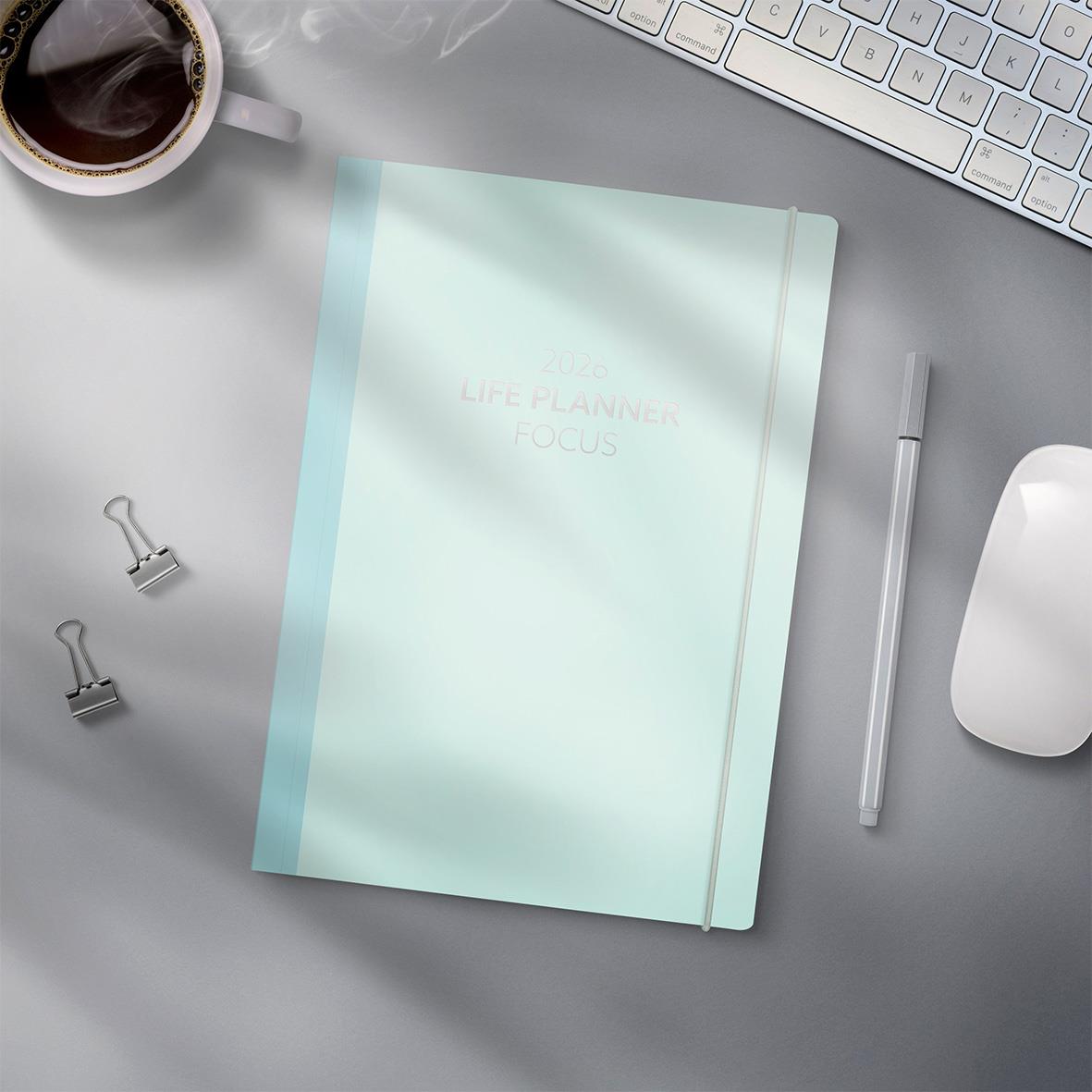 Ljusblå Life Planner Focus 2026 på skrivbord med penna, koppar kaffe, tangentbord, mus och klämmor