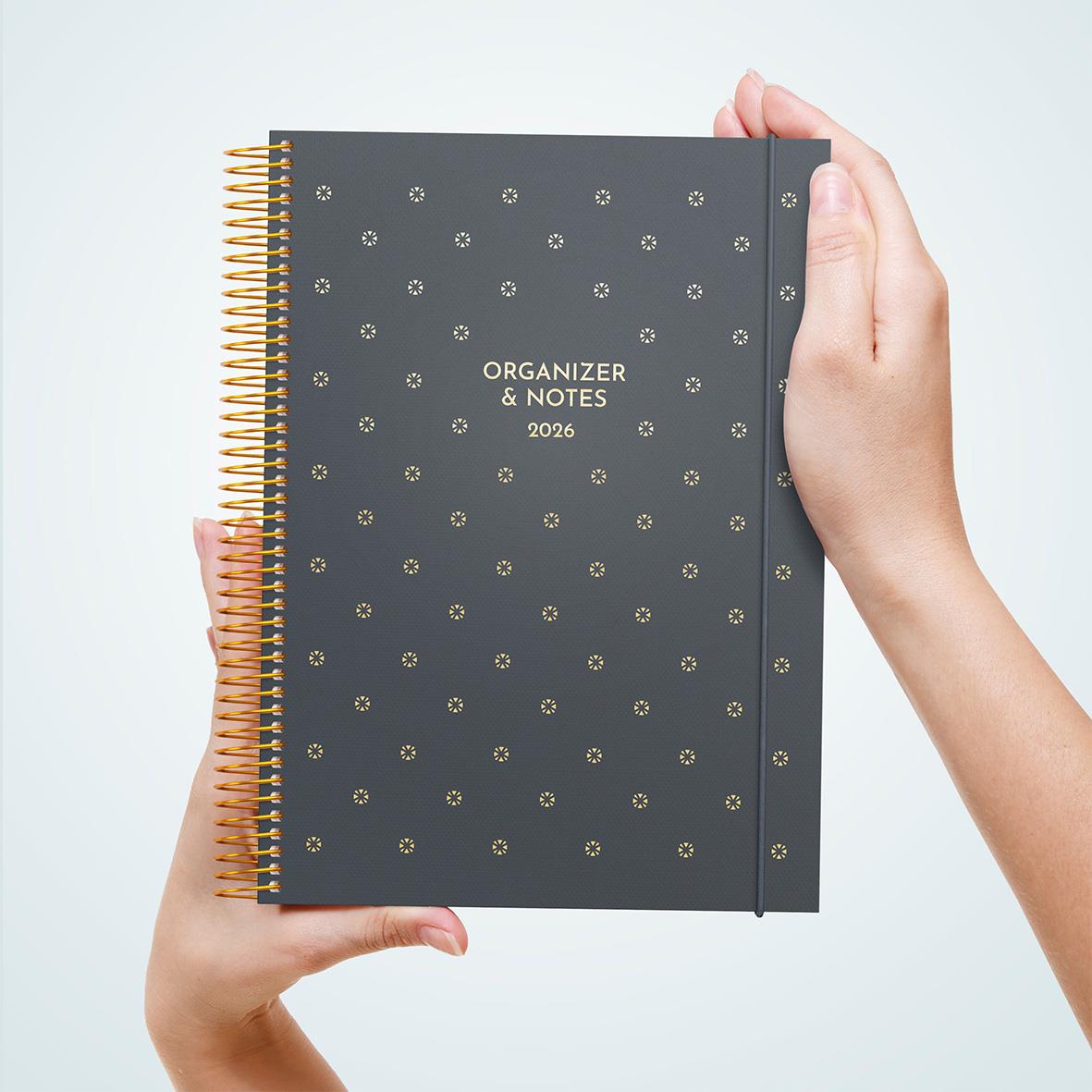 Svart almanacka med guldprickar och guldspiral, märkt Organizer & Notes 2026, hålls av en person med ljus hud