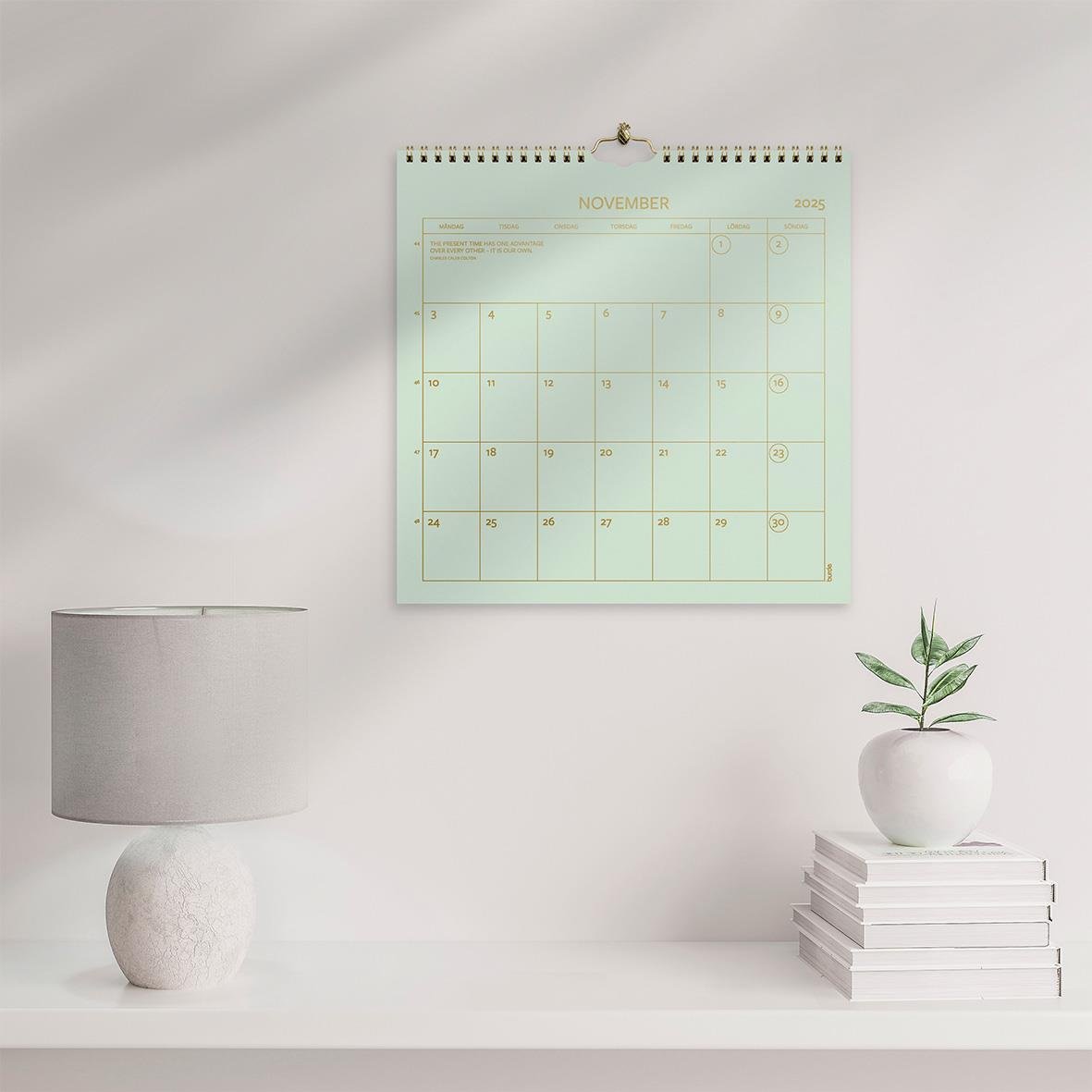 Väggkalender för november 2026 hängande på vit vägg med växt och bokstaplar bredvid