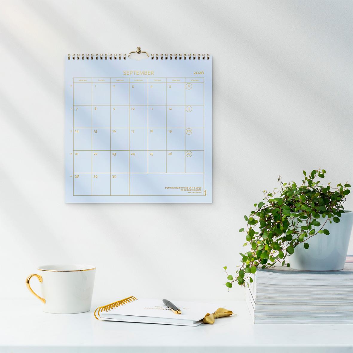 Väggkalender Burde 1752 Väggkalender Color 2026 på vit vägg med kopp, anteckningsblock och växt