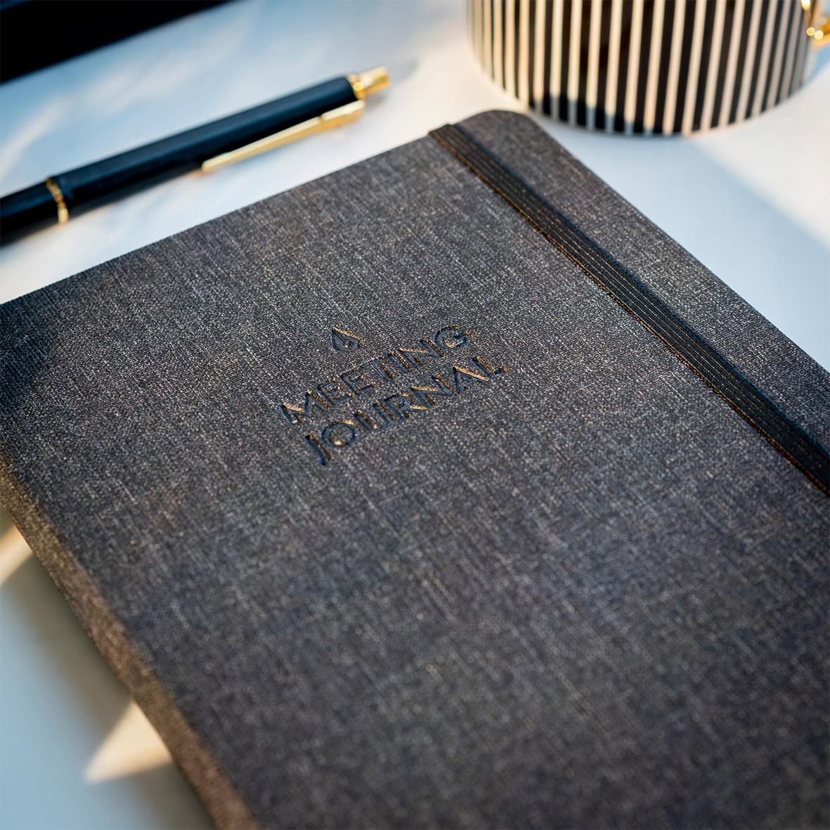Svart mötesjournal med guldtext och elastisk band, omgiven av penna och dekorativ vas