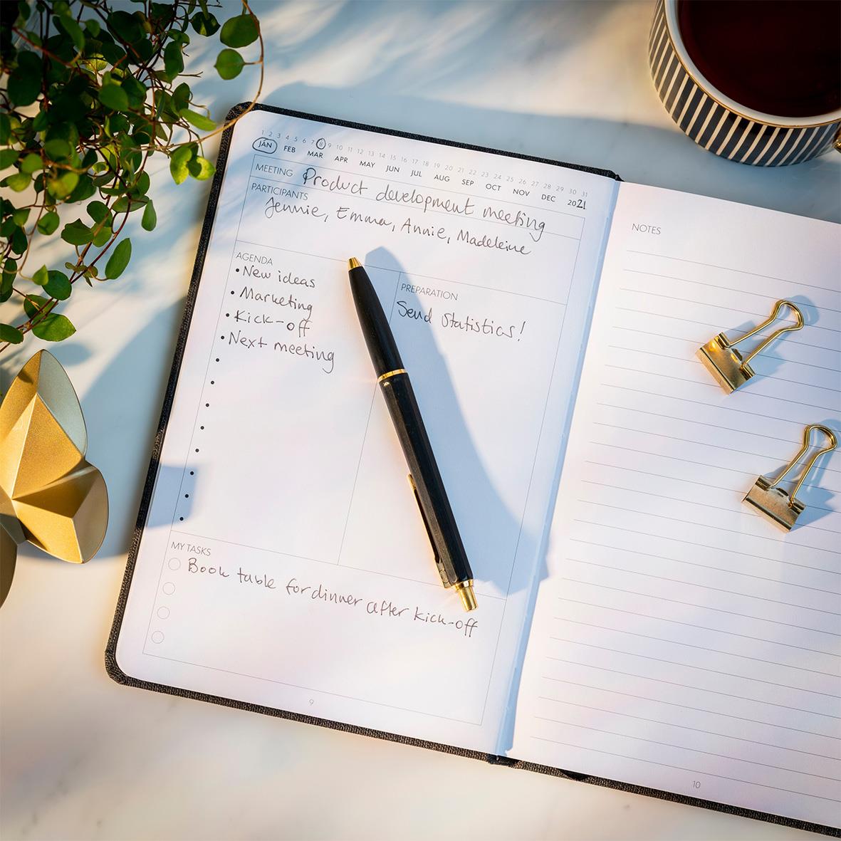 Öppnad mötesjournal med penna, guldklämmor, kopp kaffe, växt och gulddekoration på skrivbordet