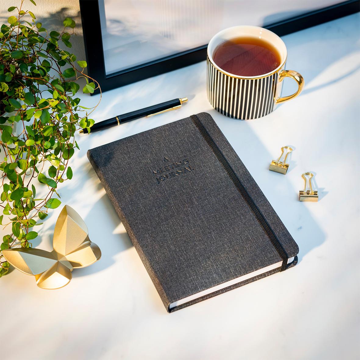 Svart anteckningsbok med elastisk band, kopp med te, guldfärgade klämmor, penna, växt och butterfly-dekoration på skrivbordet