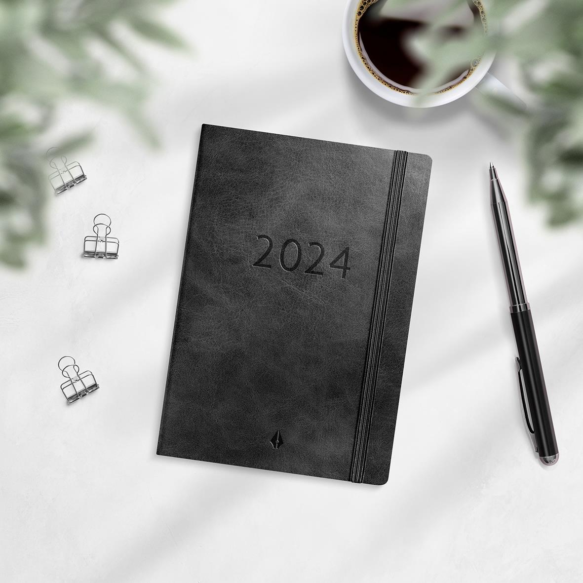 Svart 2024 års almanacka med elastisk band på vit bakgrund, omgiven av klämmor, penna och en kopp kaffe