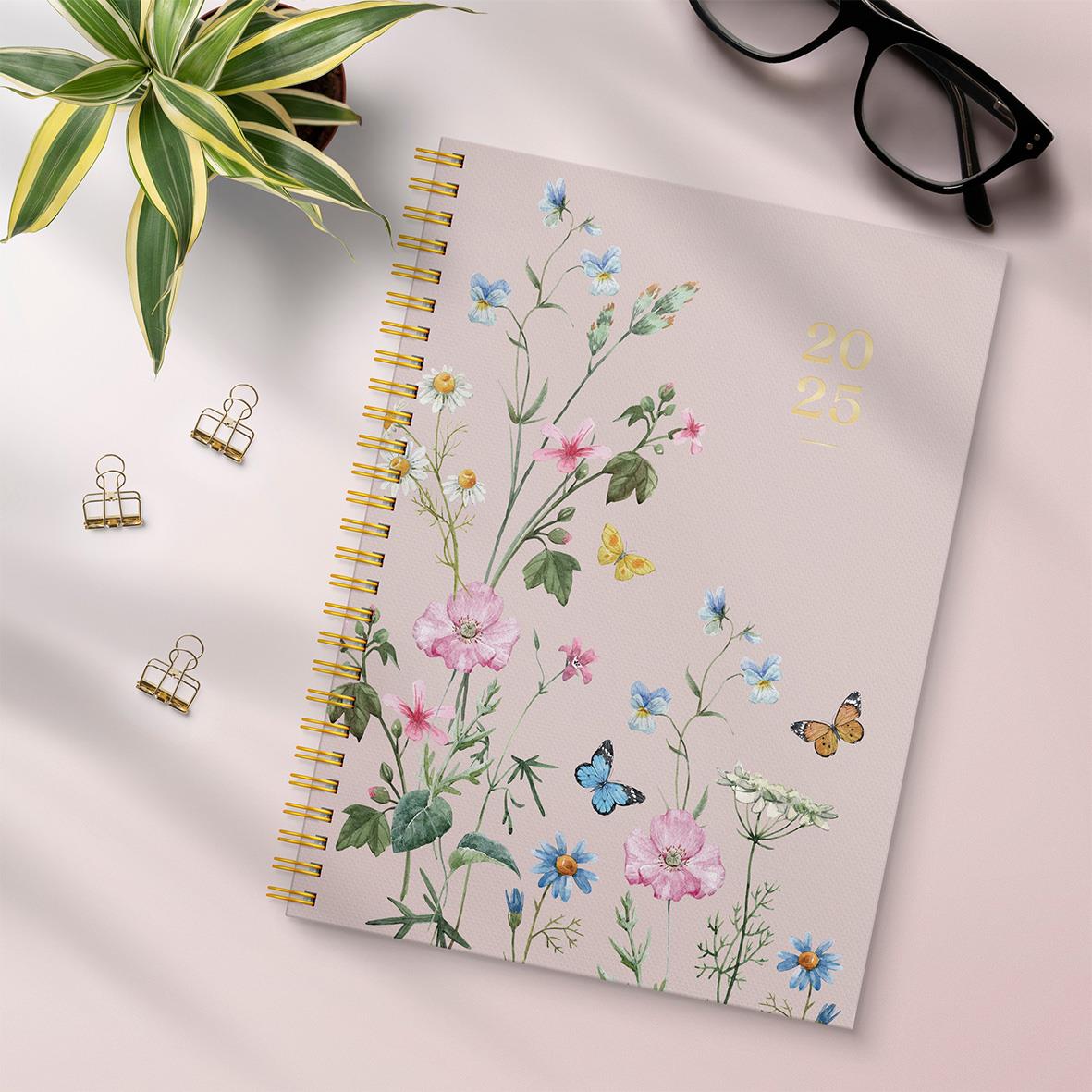 Våralmanacka 2025 med blommor och fjärilar, guldkantad spiralbindning, omgiven av gröna växter, glasögon och guldklämmor