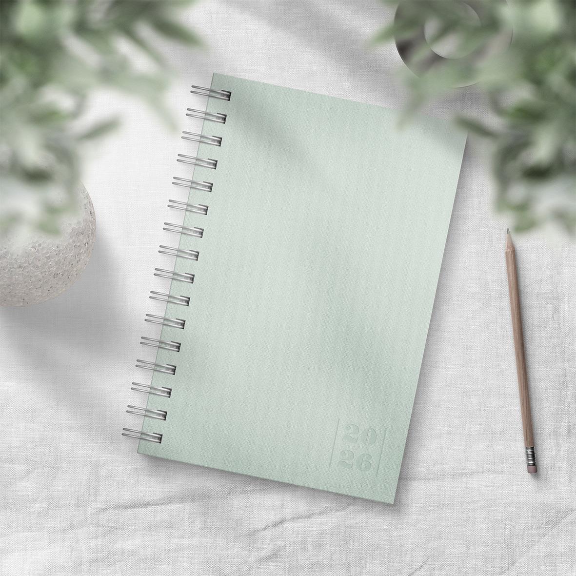 Grön dagbok med spiralbindning och penna på vit duk, omgiven av gröna växter