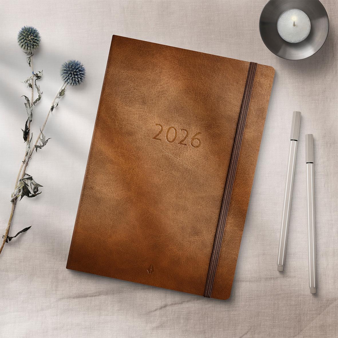 Brun 2026 års almanacka med elastisk band och enkel design, placerad på ljus bakgrund med blommor och ljus