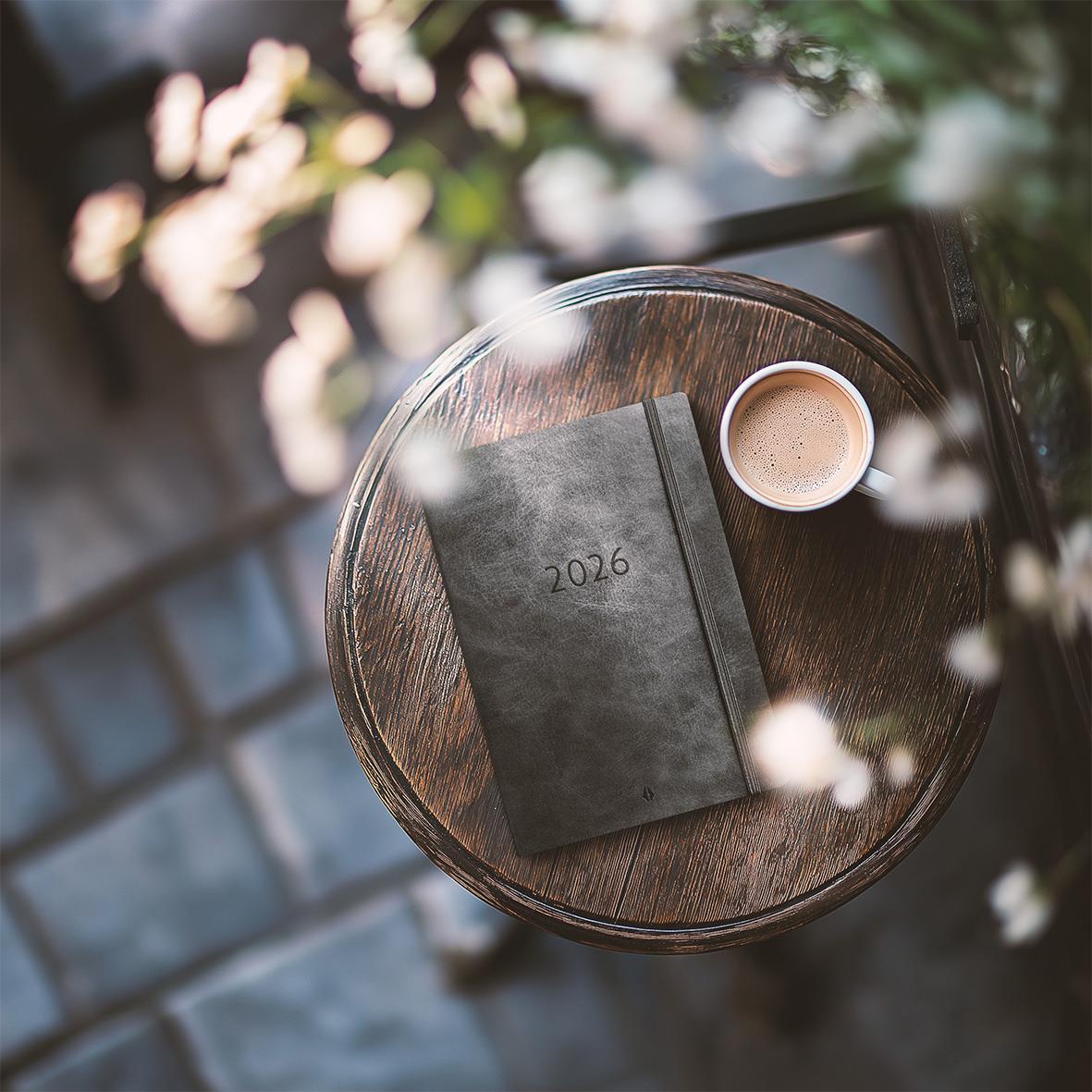 Svart Almanacka 2026 på ett runt träbord med kopp kaffe och blommor i bakgrunden