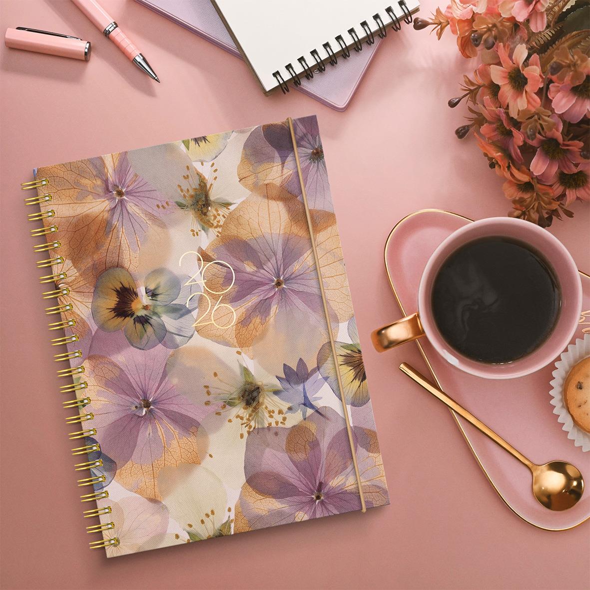 Blommig almanacka med guldspiral, penna, anteckningsblock, kopp kaffe och blommor på rosa bakgrund