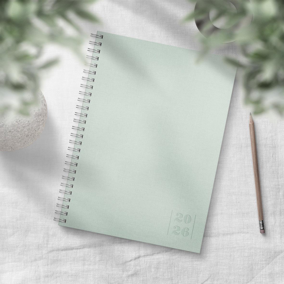 Grön almanacka för 2026 med spiralbindning och penna på vit duk, omgiven av gröna växter