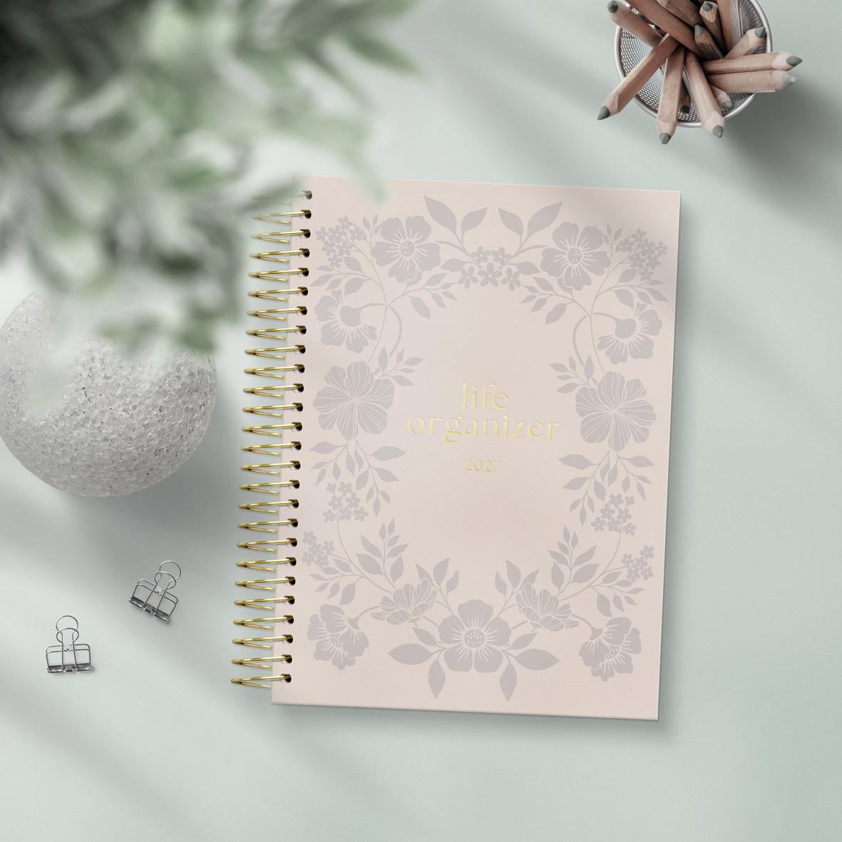 Ljusrosa A5-årskalender med blomsterdekor och guldfärgad spiralbindning, omgiven av gröna växter och kontorsmaterial