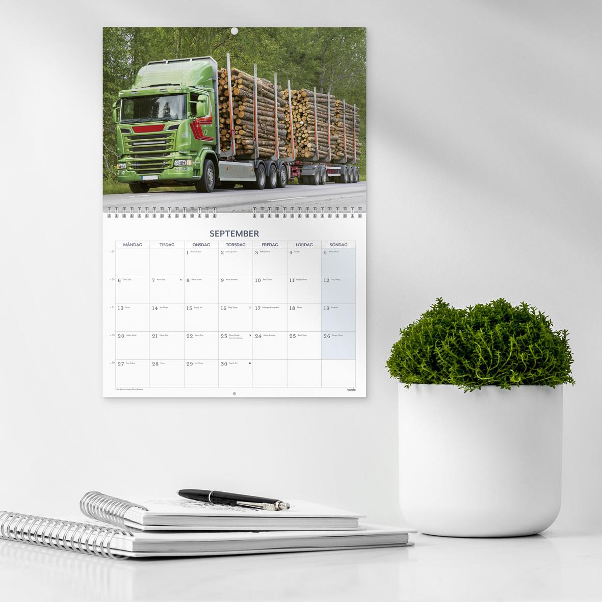 Väggkalender för september 2027 med lastbil och trästockar, placerad på en vit vägg bredvid en grön växt i kruka.