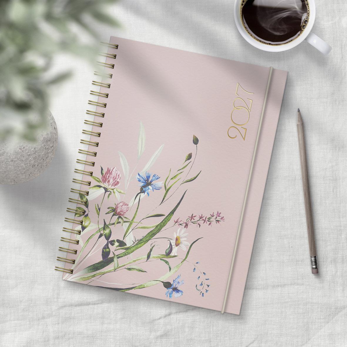 Årsalmanacka för 2027 med blommönster och spiralbindning, rosa färg, A5-storlek, placerad på ett bord med penna och kopp kaffe