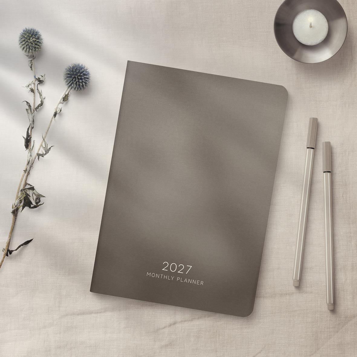 Månadsplanerare för 2027 i A5-format med grått omslag och penna, placerad på ljus bakgrund med blomma och ljus.