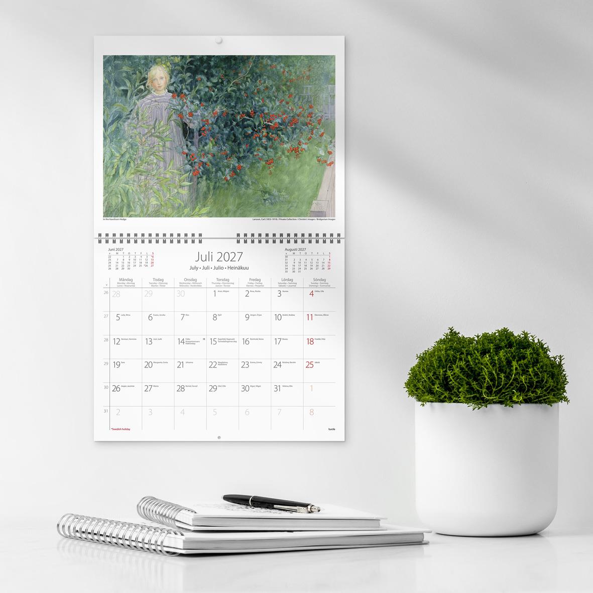 Väggkalender med Carl Larssons konst från 1741, visar juli 2027, på vit vägg bredvid grönt växt i kruka och skrivbord med anteckningsblock och penna