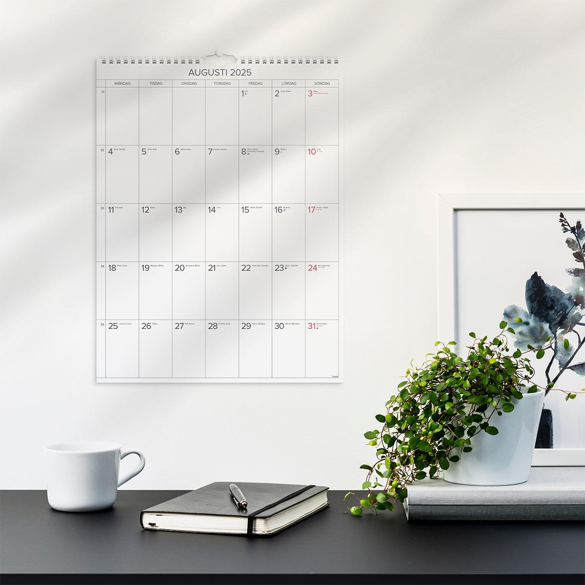 Väggkalender för augusti 2025 på vit vägg med gröna växter och skrivbord med kopp och anteckningsbok