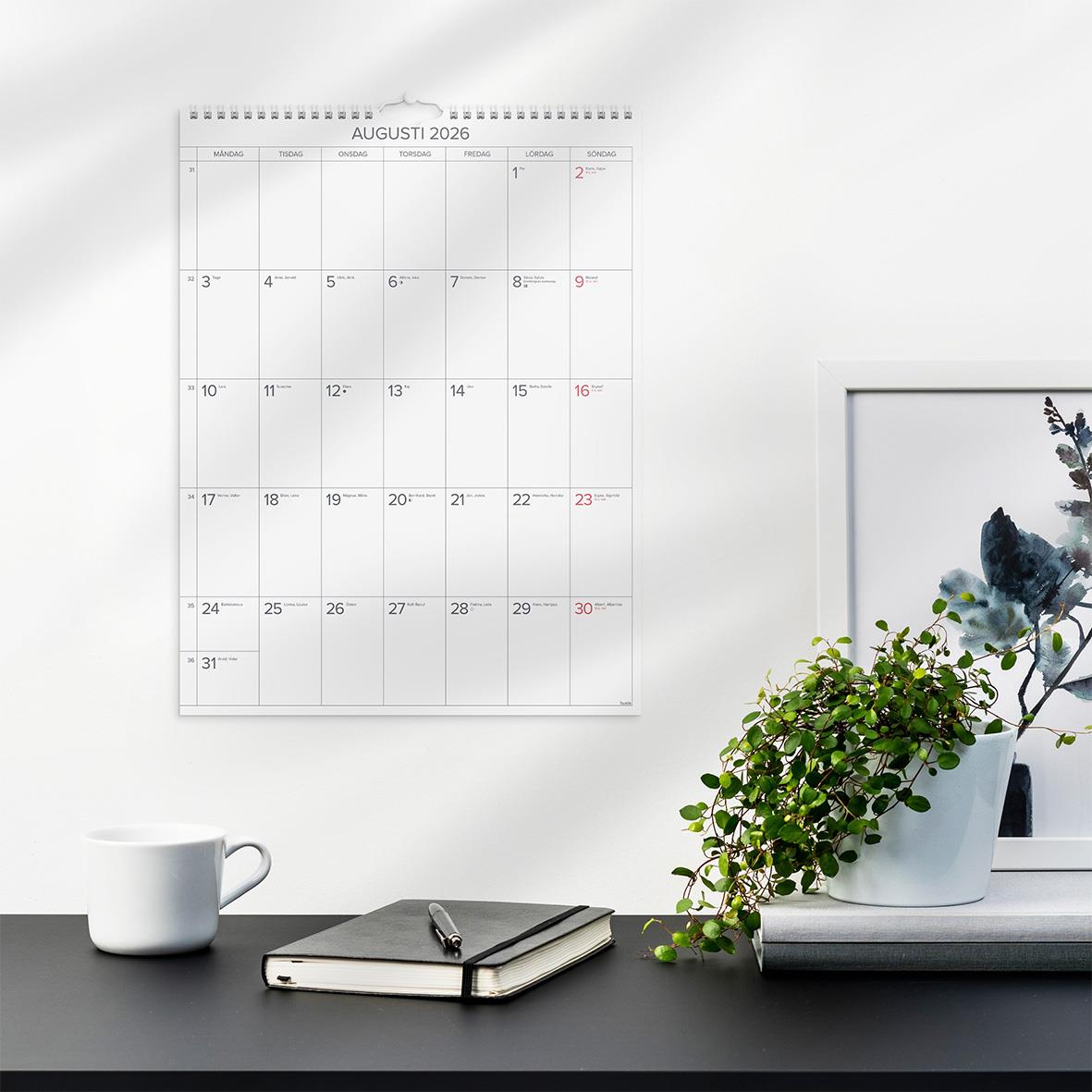 Väggkalender för augusti 2026 med stora månadsdatum, placerad på en vit vägg bredvid en växt, en kopp och en anteckningsbok.