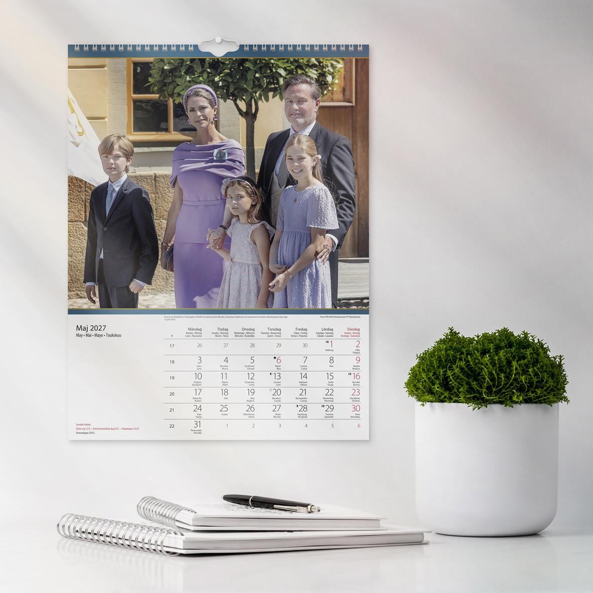 Väggkalender för 2026 med Kungafamiljen i formell klädsel, inomhusmiljö, gul soffa och konst på väggarna