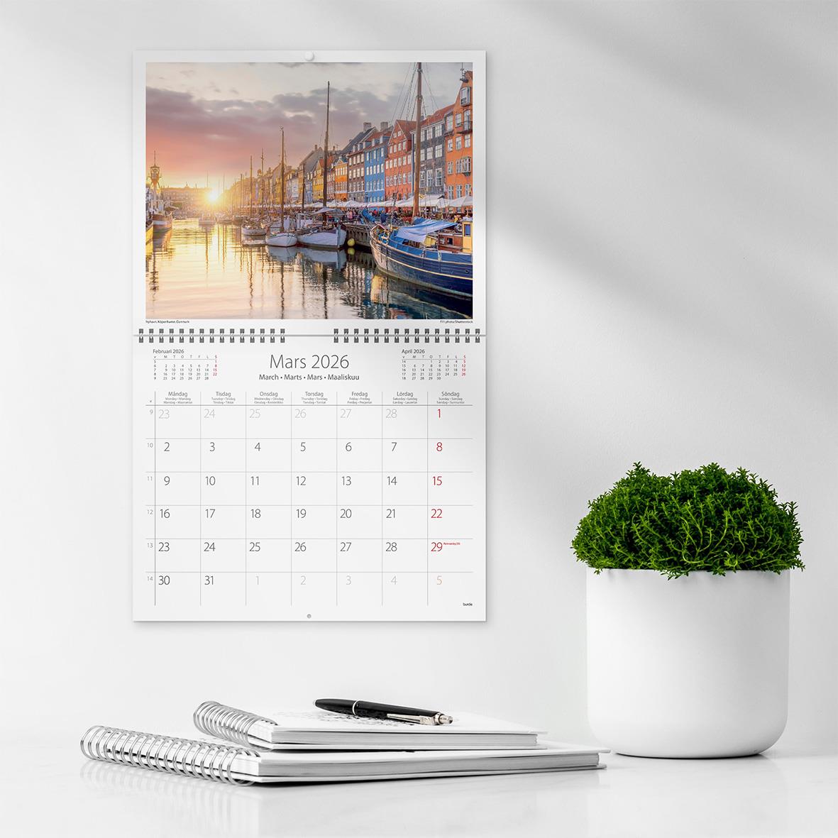 Väggkalender för mars 2026 med bild av en hamn vid solnedgång, på en vit vägg bredvid en grön växt och skrivbord