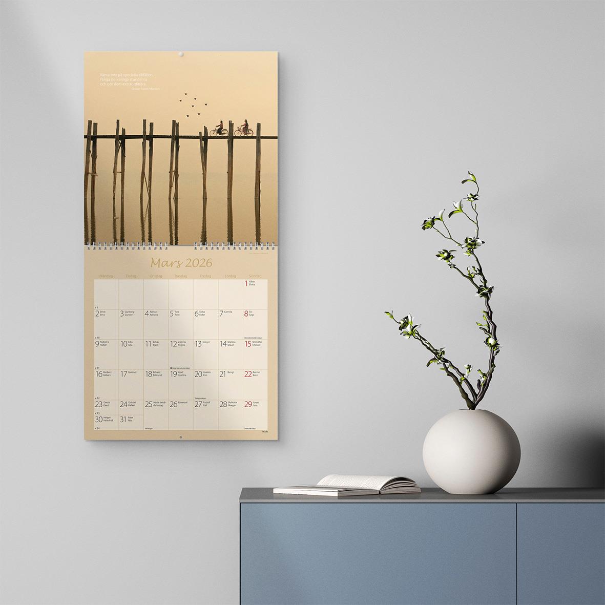 Väggkalender Burde 1721 Carpe Diem 2026 med bild av en brygga och fåglar, samt en vit vas med en gren på en blå möbel