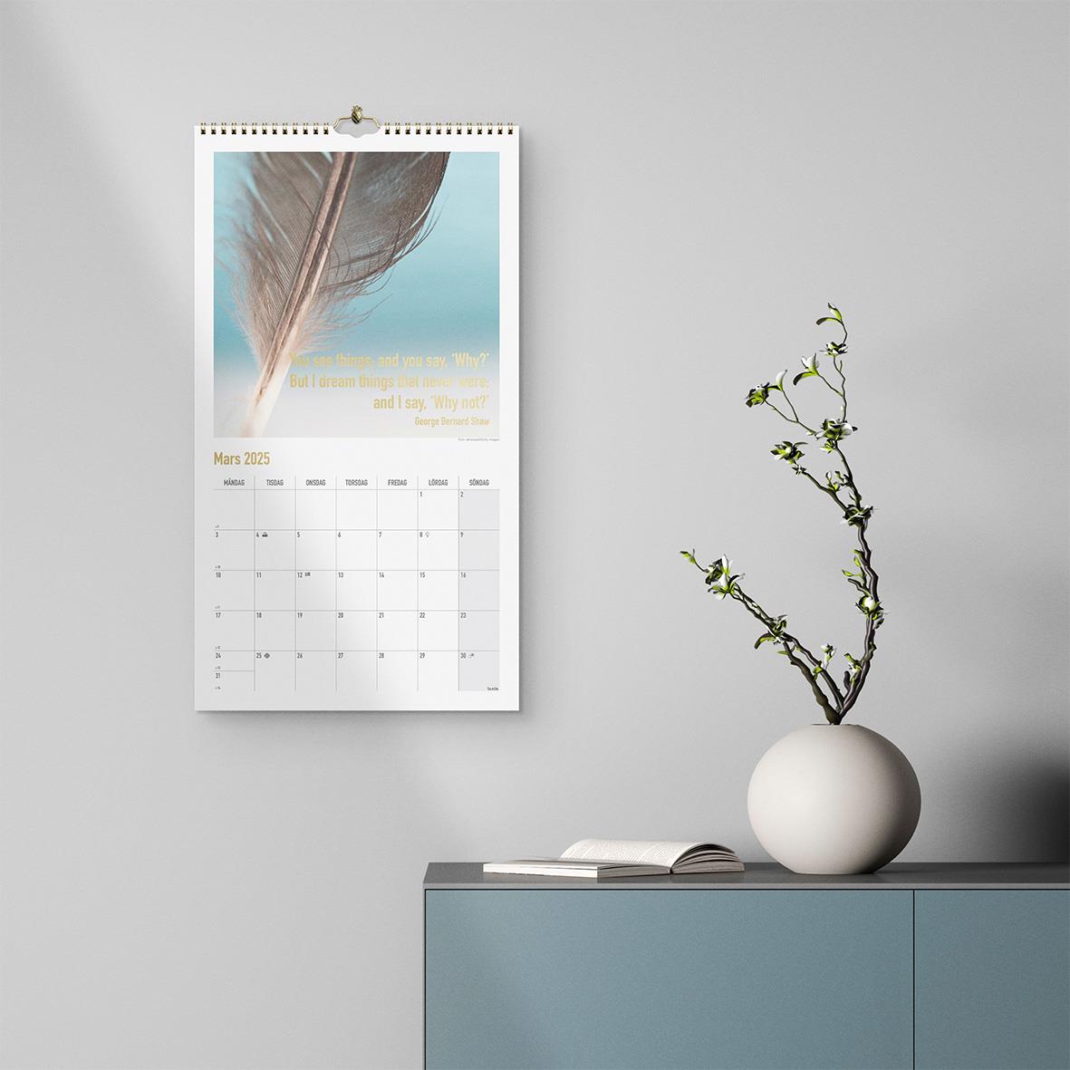Väggkalender Burde 1736 Dream & Do 2026 hänger på en vägg bredvid ett blått skåp med en vit vas och en växt