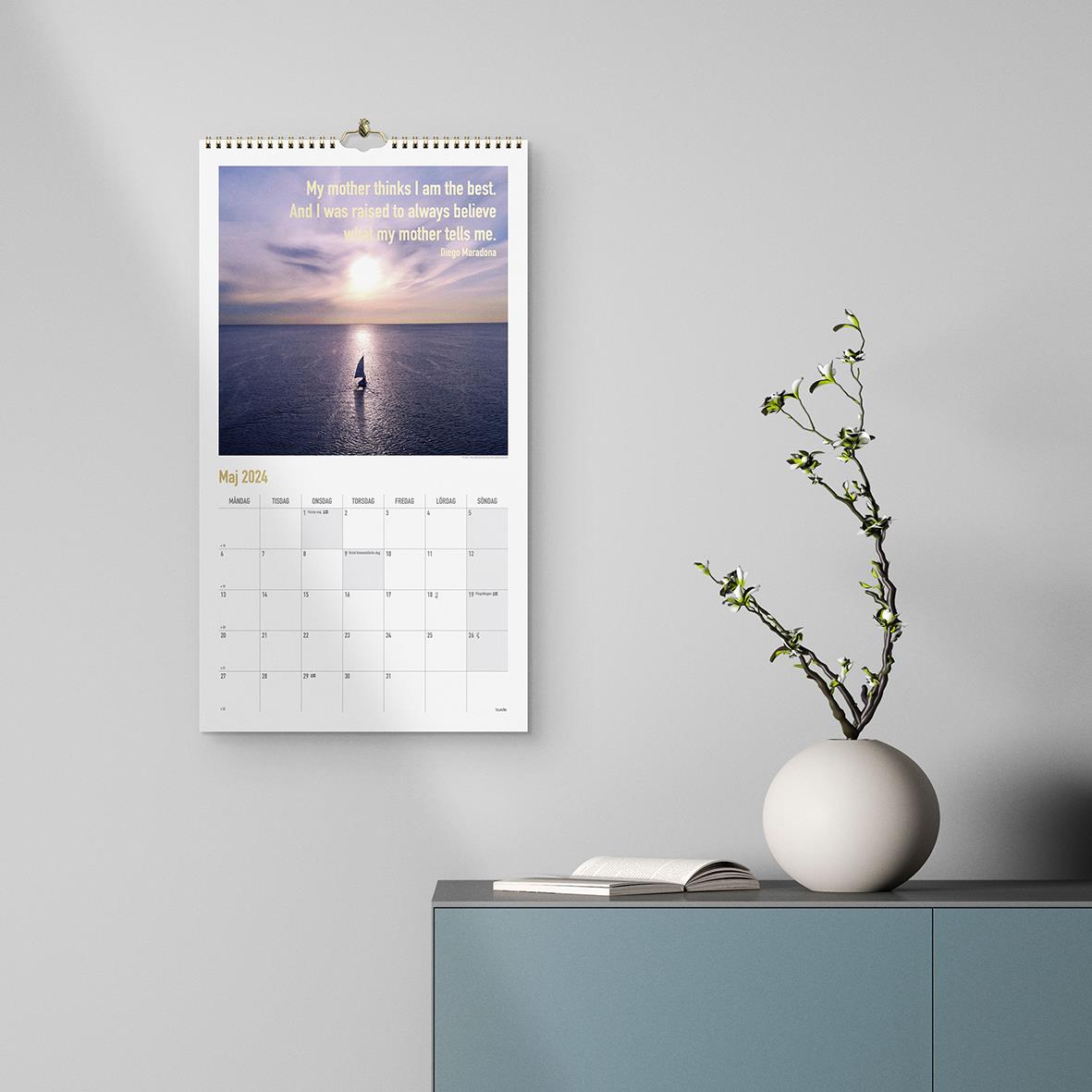 Väggkalender Burde 1736 Dream & Do 2026 med bild av hav och solnedgång, hängande på vägg bredvid en växt i vas