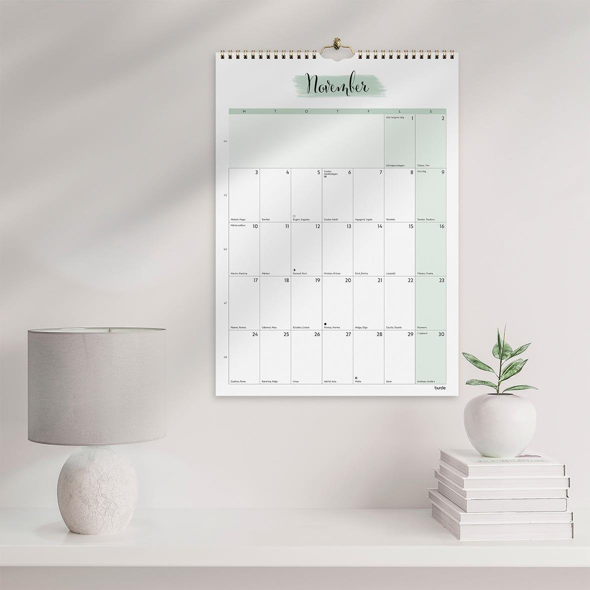 Väggkalender för november 2026 med plats för anteckningar, hängande på en vägg bredvid en lampa och en växt