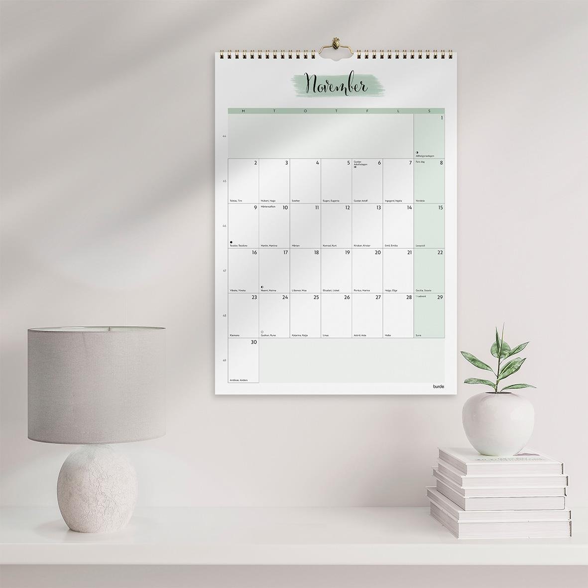 Väggkalender för november 2026 med plats för anteckningar, hängande på en vägg över ett bord med en lampa och växt
