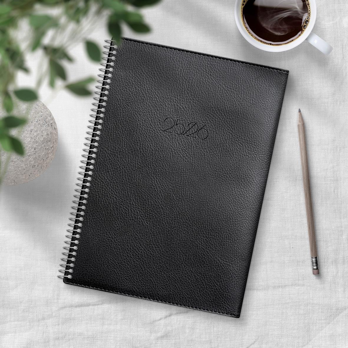 Svart A5 studiealmanacka med spiralrygg och embossed märke, omgiven av en kopp kaffe, penna och gröna växter