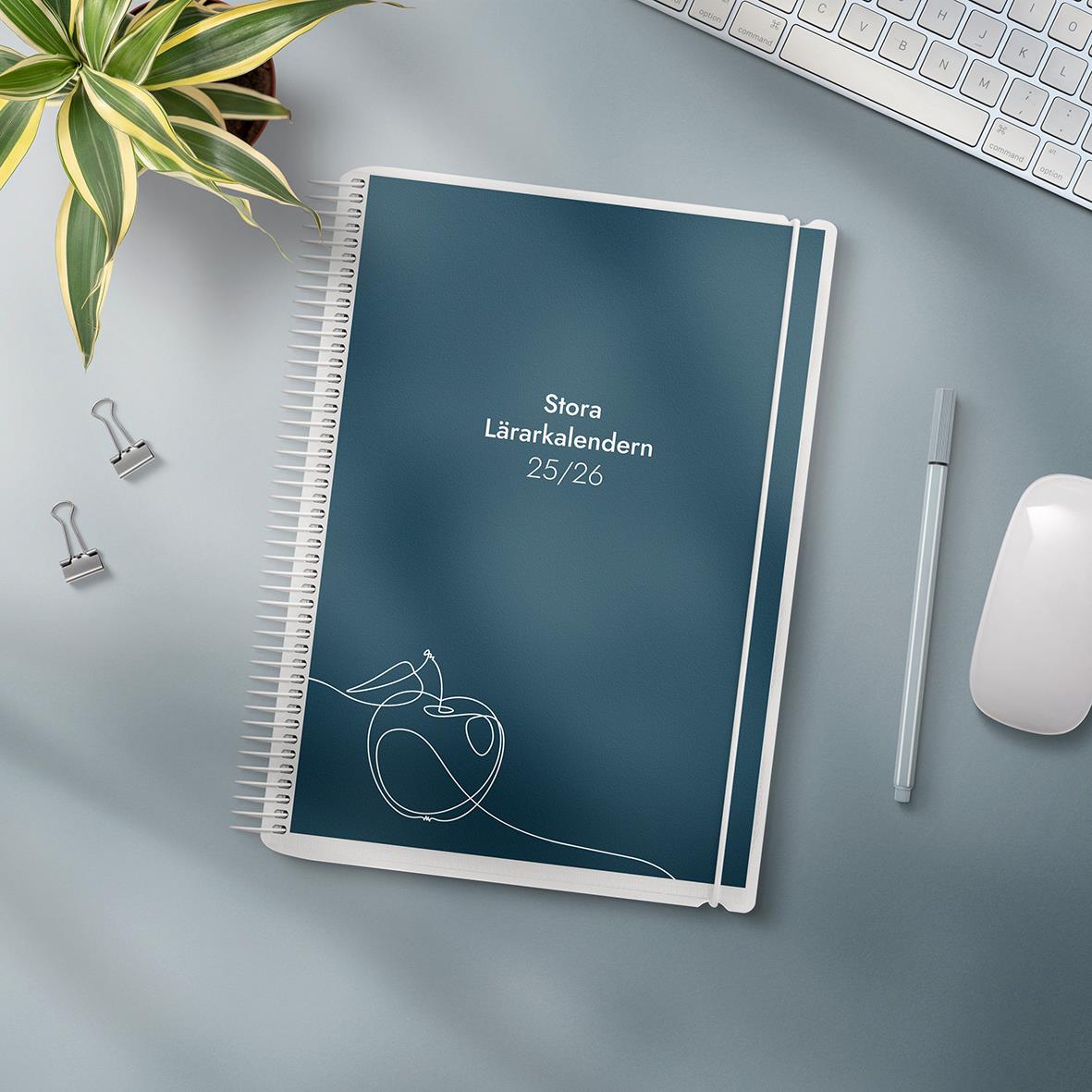 Stort lärarekalender för 2025/2026 med spiralbindning och blå omslag, omgiven av växt, tangentbord, penna, och mus på skrivbord