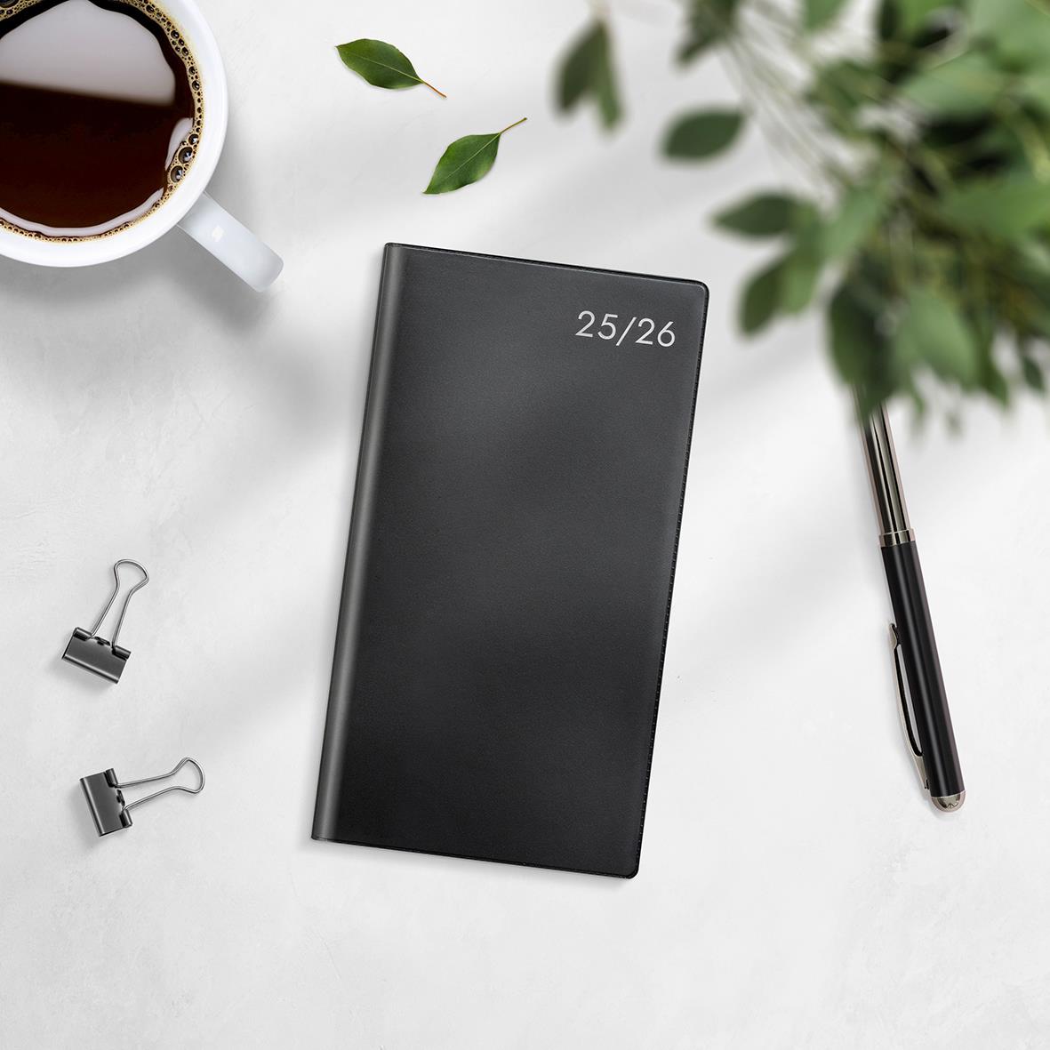 Svart studiealmanacka för 2025/26 med titeln Stora Basic Plast 3681, omgiven av penna, klämmor, växt och kopp kaffe