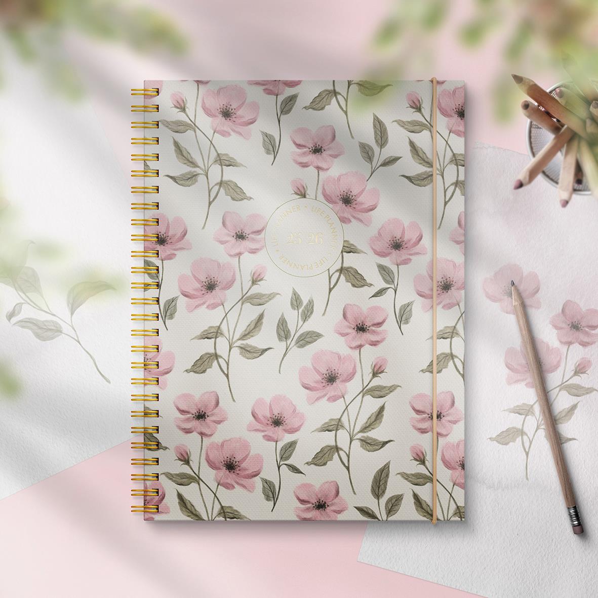 A5 livsplanerare med blommönster i rosa och grönt, spiralbindning, på ljus bakgrund med pennor och papper