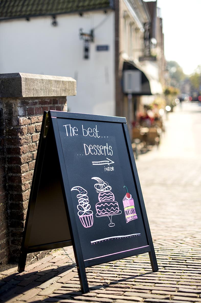 Gatuställ Securit Woody Furu svart 47x67cm med skrivyta, utomhus på trottoaren, visar en svart anslagstavla med teckningar av desserter