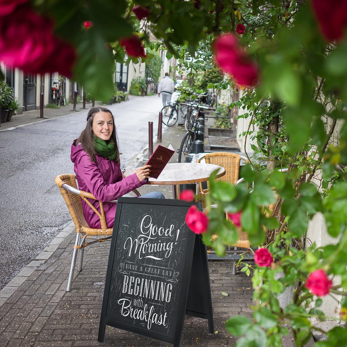 Kvinna sitter vid uteservering med svart skylt med text Good Morning och Begin with Breakfast, omgiven av rosa blommor