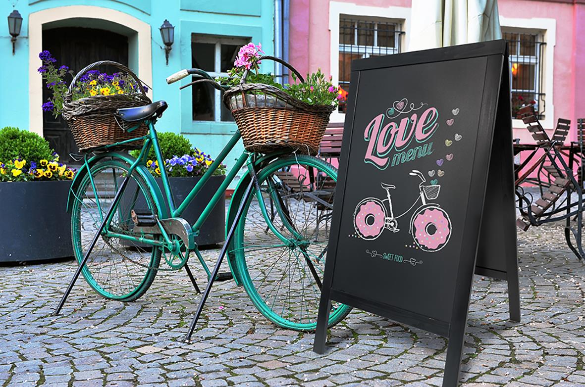 " Securit Woody Furu svart 58x87cm med skrivyta, cykel och blomkrans framför färgglada hus"