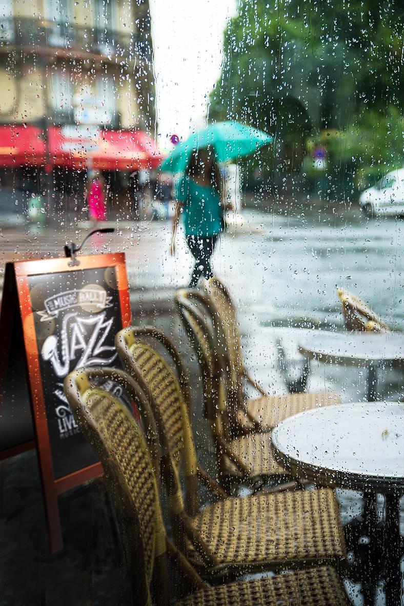 Utsikt genom ett fönster med regndroppar, uteservering med stolar och bord, person med paraply i regnet