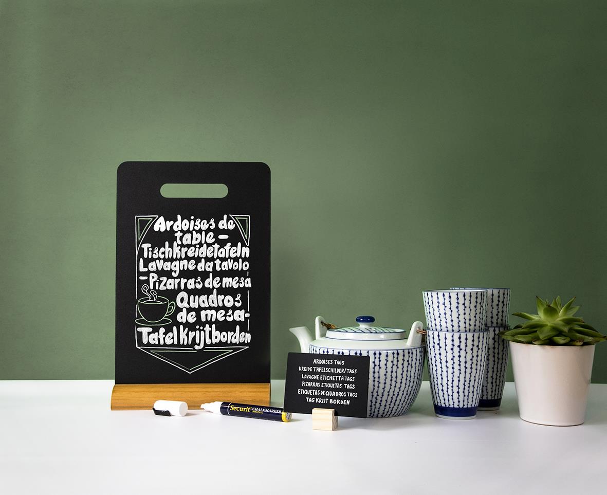 Svart griffeltavla med handtag, markerpen, och växt på bord mot grön vägg