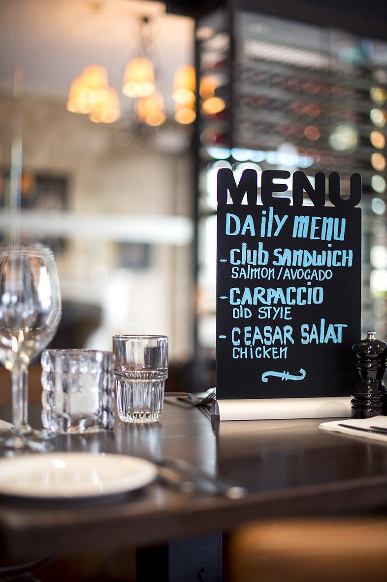 Svart griffeltavla med meny för restaurang, markerad med märkpennor, på bord med glas och tallrik i bakgrunden
