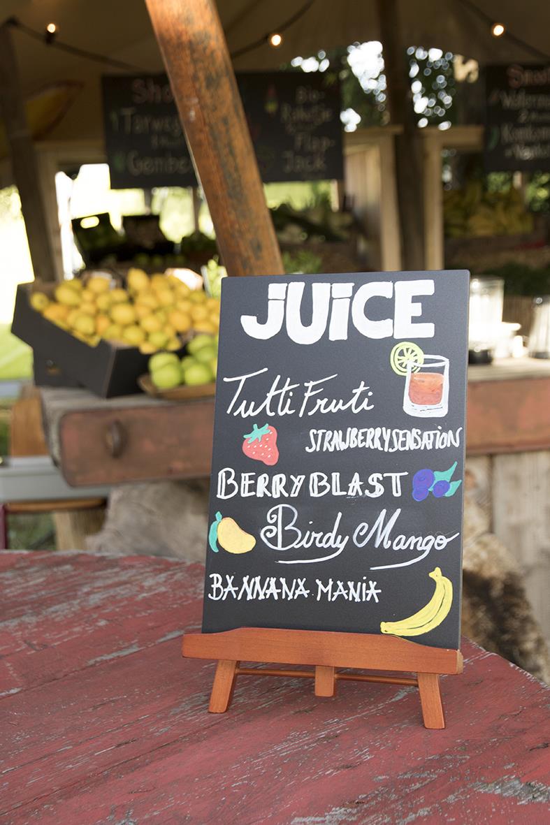 Svart griffeltavla med handskriven meny för juice och frukt, dekorerad med färgglada fruktillustrationer, på ett trädgårdsbord