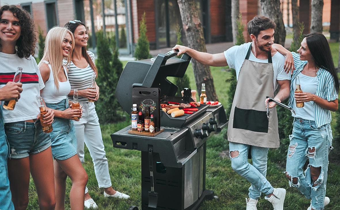 Grillfest med fyra personer som grillar och dricker utomhus, med en svart grill och ett griffeltavla bordscaddy