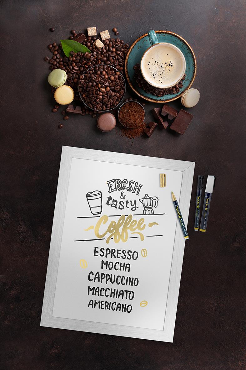 Vit griffeltavla med svart text och illustrationer, omgiven av kaffe, choklad och skrivmaterial på mörk bakgrund