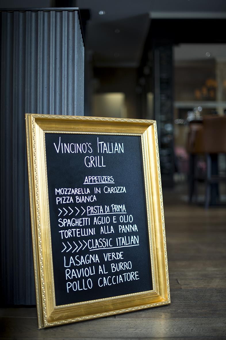 Svart griffeltavla med guldram och italienska maträtter skrivna med vit krita, placerad på golvet i ett inomhusmiljö