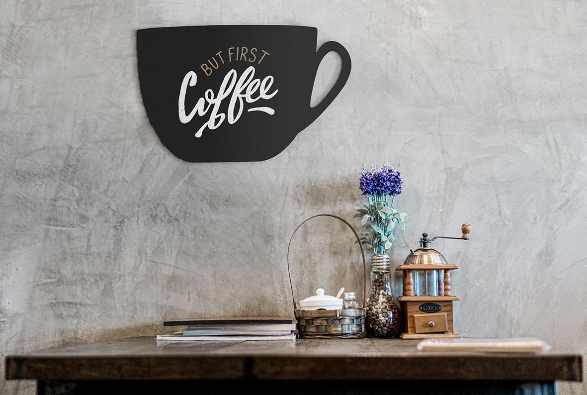 Svart griffeltavla i form av kaffekopp med texten But First Coffee på vägg ovanför ett bord med blommor och kaffekvarn