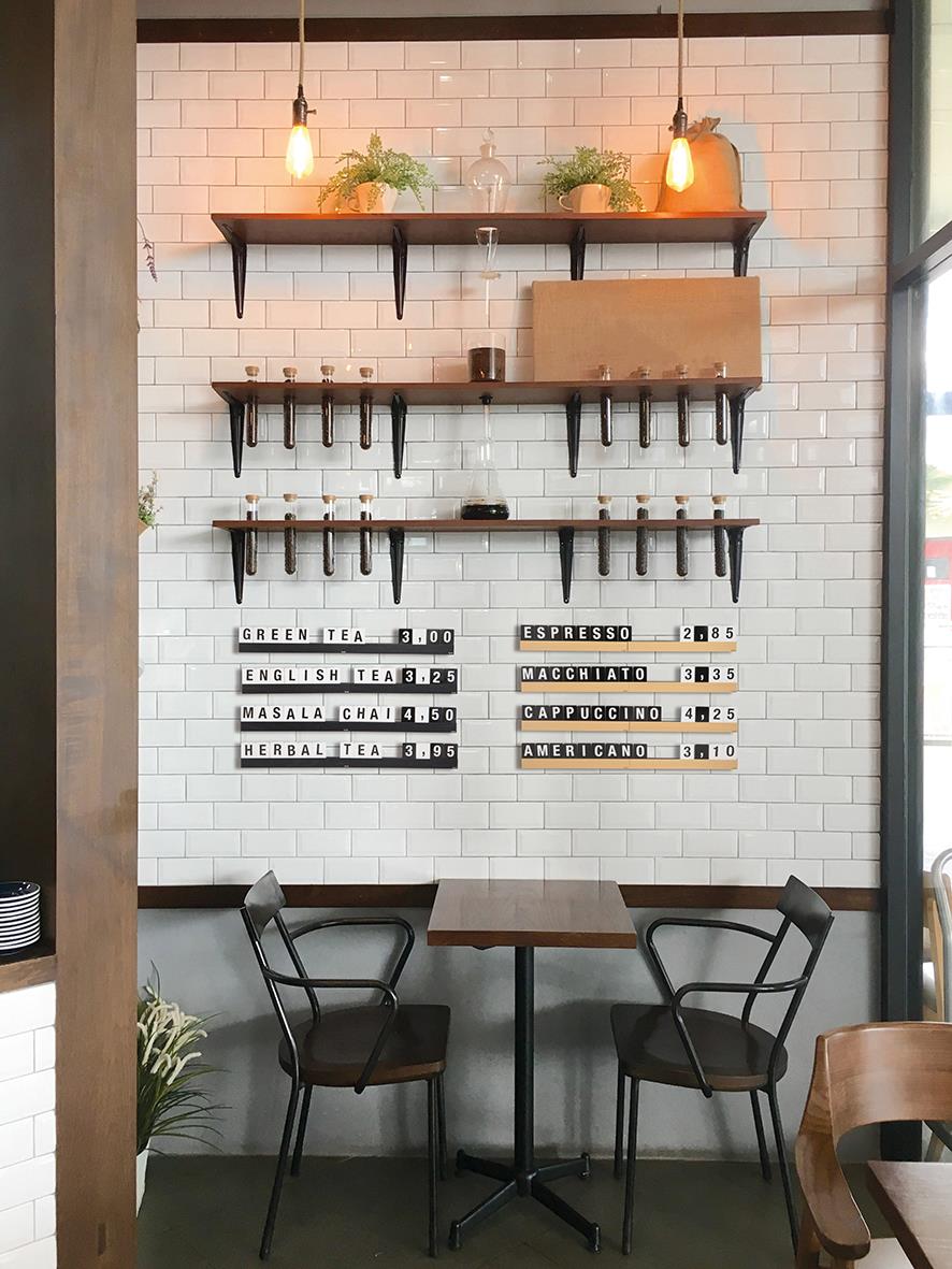 Svart bokstavshylla med tre hyllor och två lampor i ett café, vita kaklade väggar och två stolar framför ett bord.