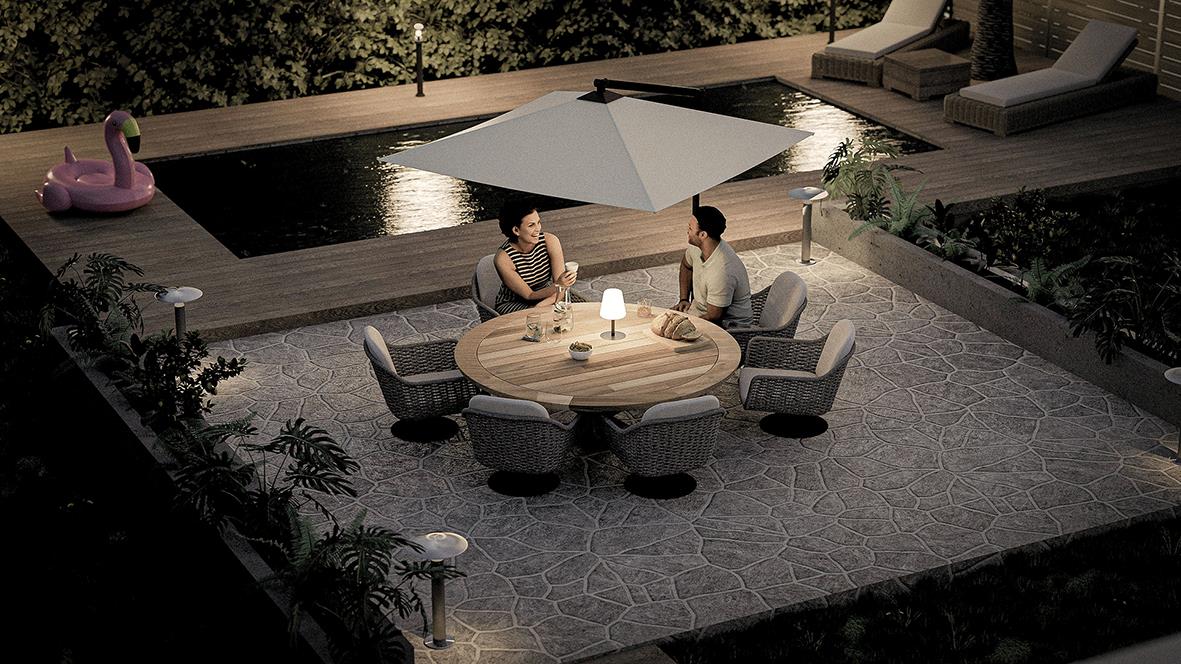 Två personer sitter vid ett runt bord med bordslampa och parasoll vid en pool på kvällen