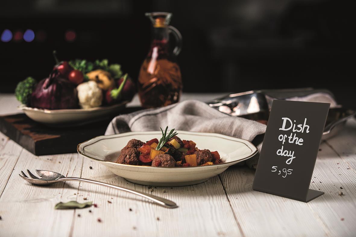 Svart griffeltavla i L-form av akryl med text dish of the day 5,95 kr på ett bord med mat och bestick